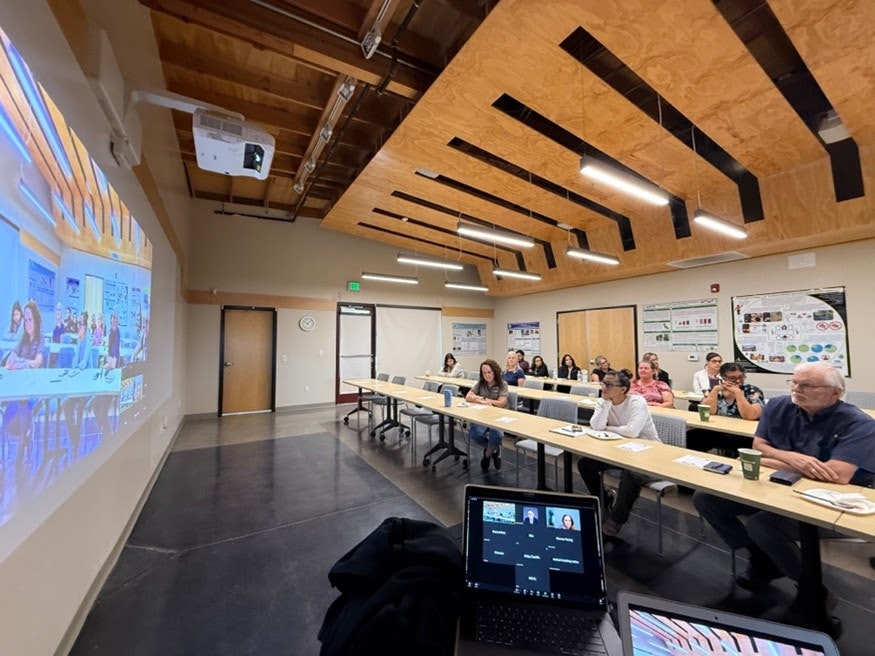 A group of people in a meeting room watching a presentation with a screen projected on the wall and participants connected on a laptop.