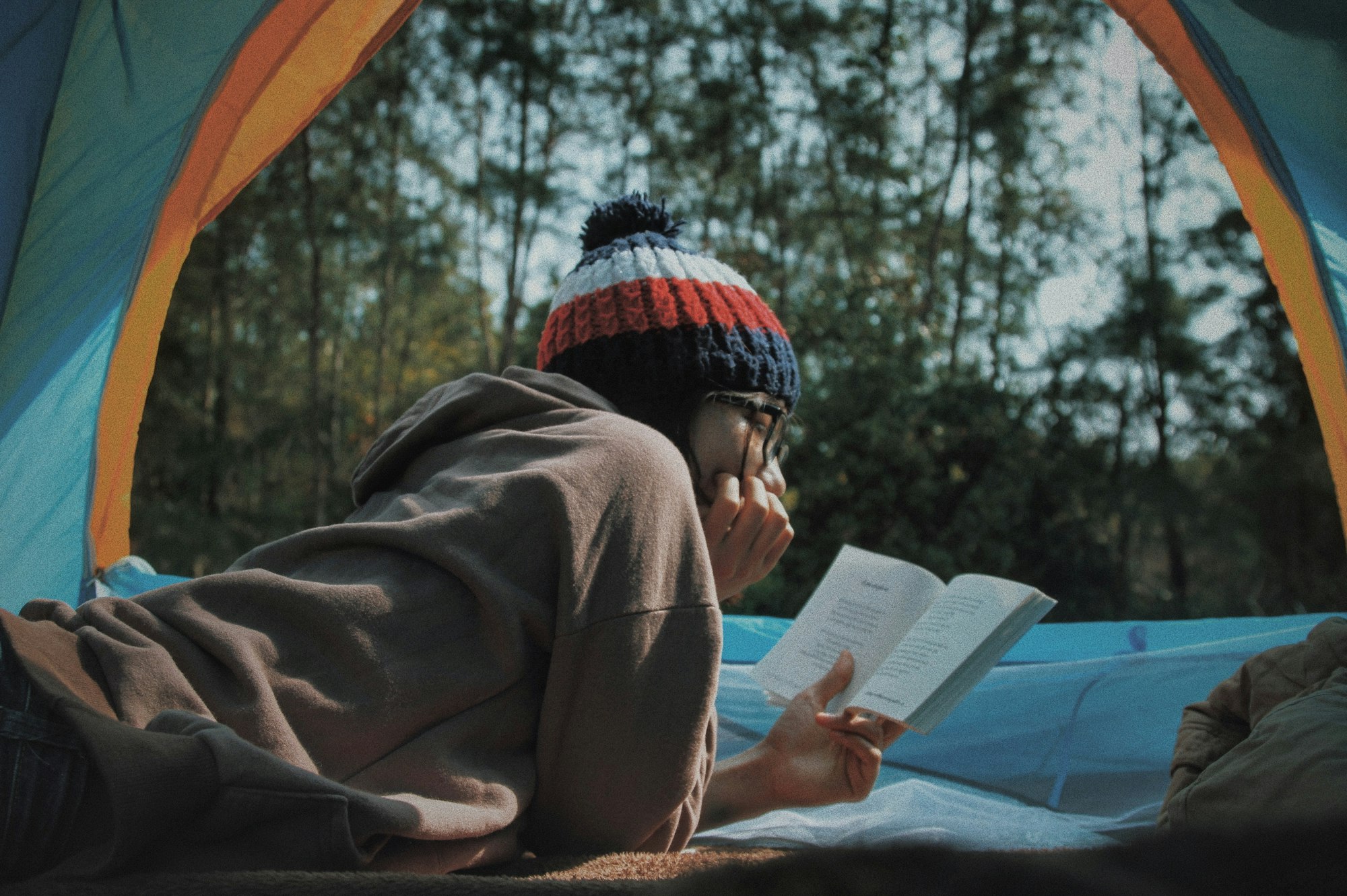 A person lies in a tent, wearing a colorful beanie, reading a book peacefully in nature.
