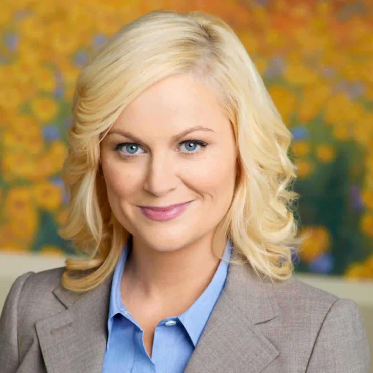 A woman with blonde, wavy hair, wearing a blazer, smiles against a colorful floral background.