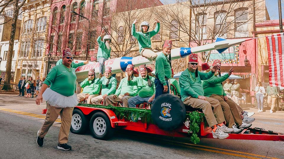 A group of people in festive attire ride on a colorful parade float shaped like a rocket, with buildings and spectators in the background.