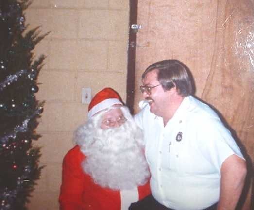 A man in glasses smiles with someone dressed as Santa Claus, near a Christmas tree and a wooden door.