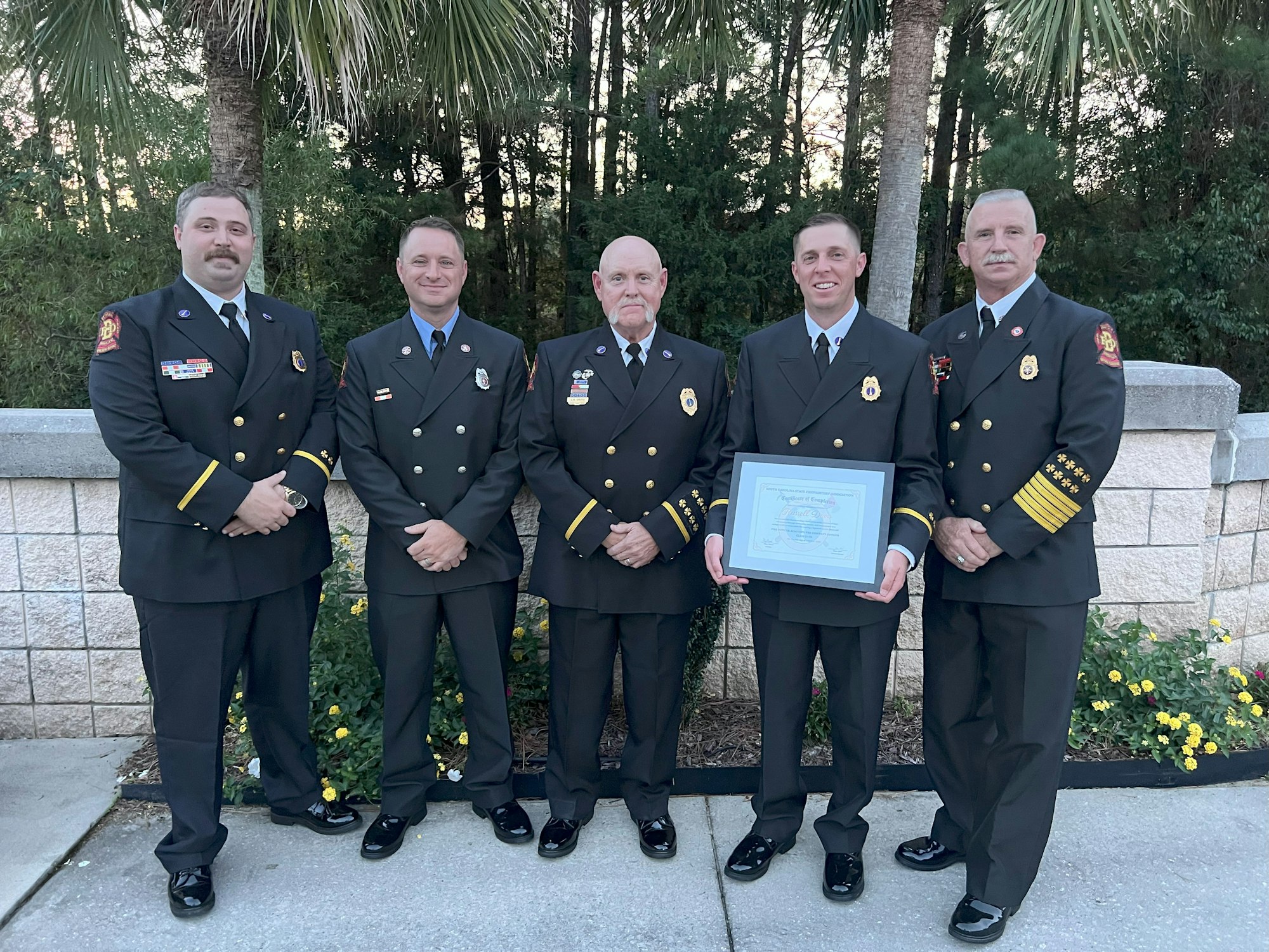 Five men in uniform, one holding a framed certificate, standing in front of trees and a wall.