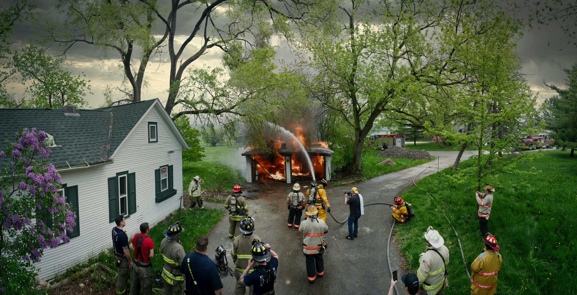 Firefighters tackling a blaze in a small building, with onlookers and emergency vehicles present.