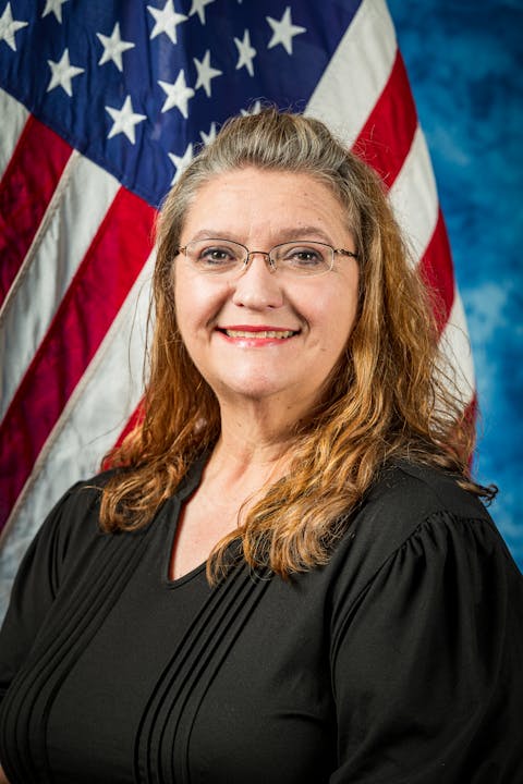 A person with glasses stands smiling in front of an American flag backdrop.
