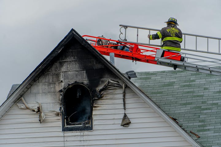 Firefighter on a ladder next to a charred attic window of a house after a fire.