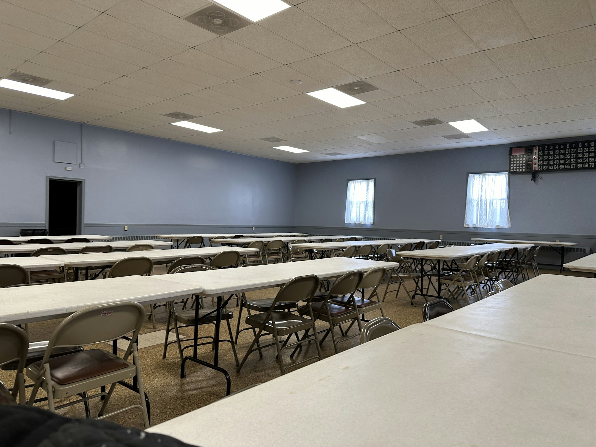 A vacant room with rows of tables and chairs, a calendar on the wall, and windows with curtains.