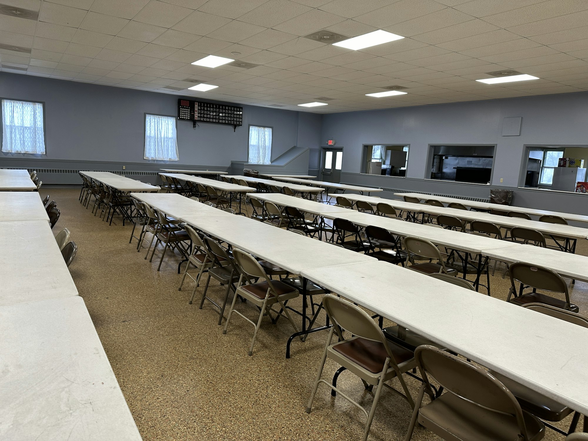 Empty dining hall with rows of tables and chairs, a service window, and a basketball scoreboard on the wall.