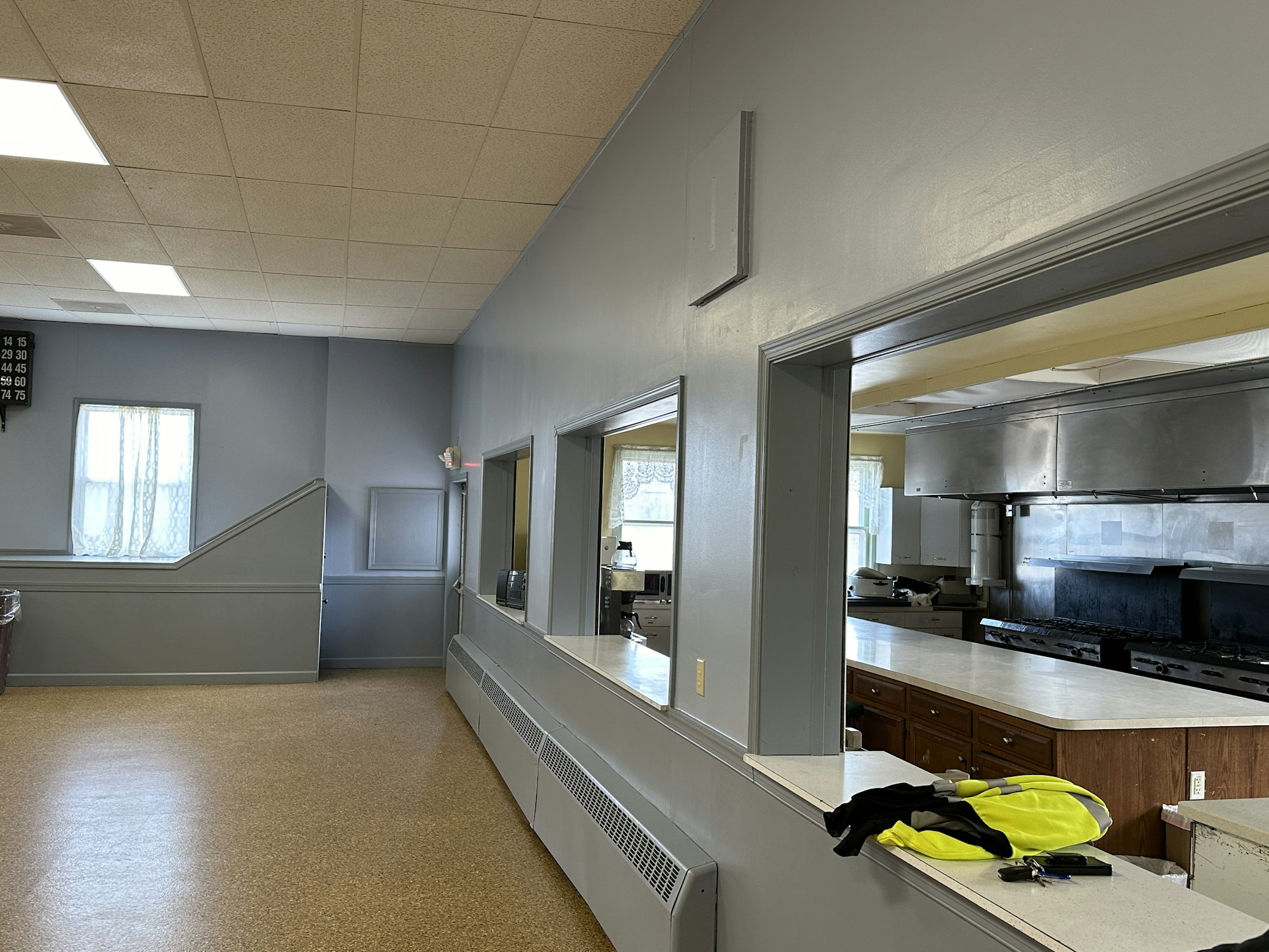 An empty commercial kitchen viewed from a serving window, with stainless steel appliances and a bingo number board on the wall.