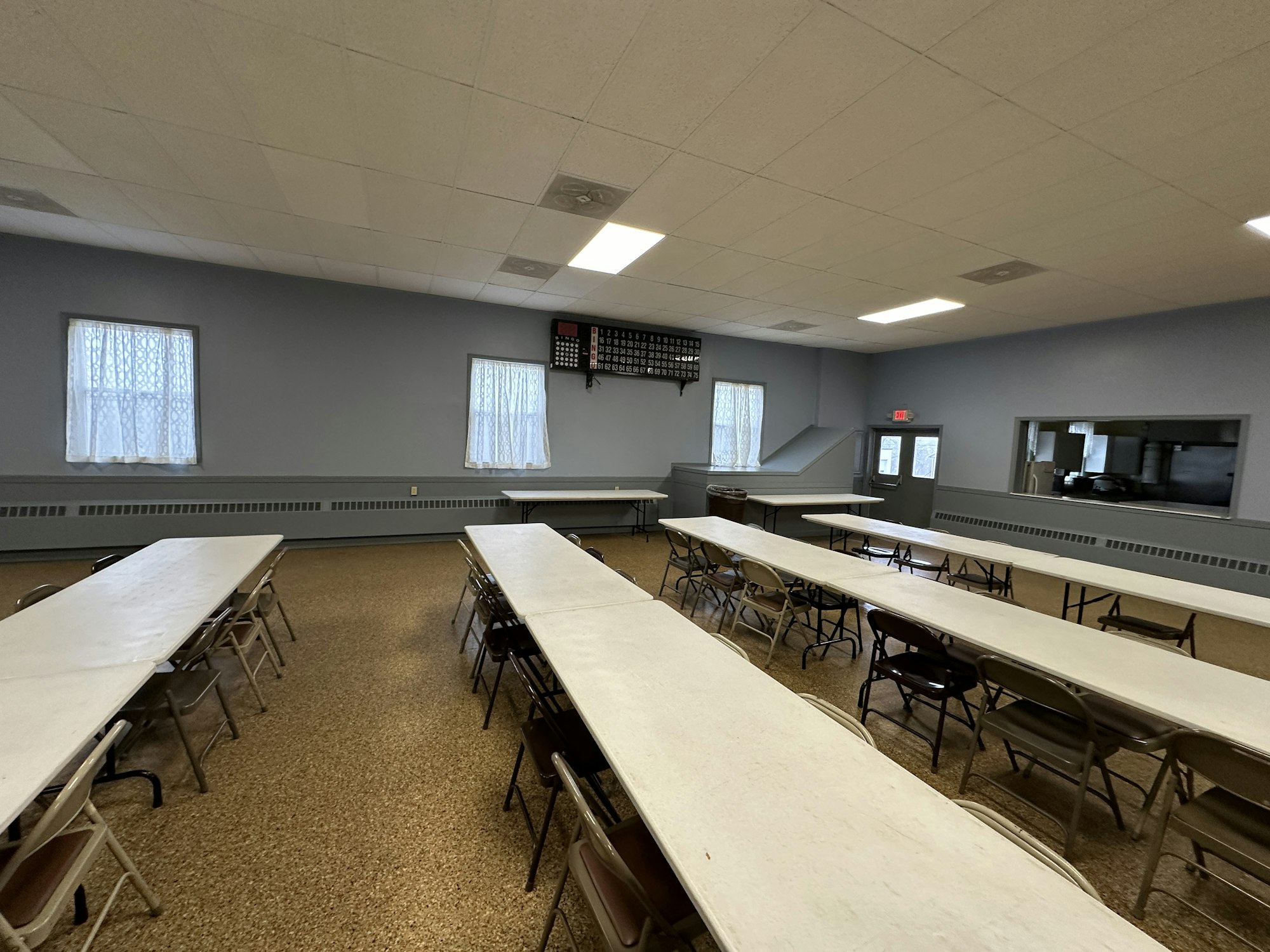 A sparsely furnished room with folding tables, chairs, a serving window, and a calendar clock on the wall.