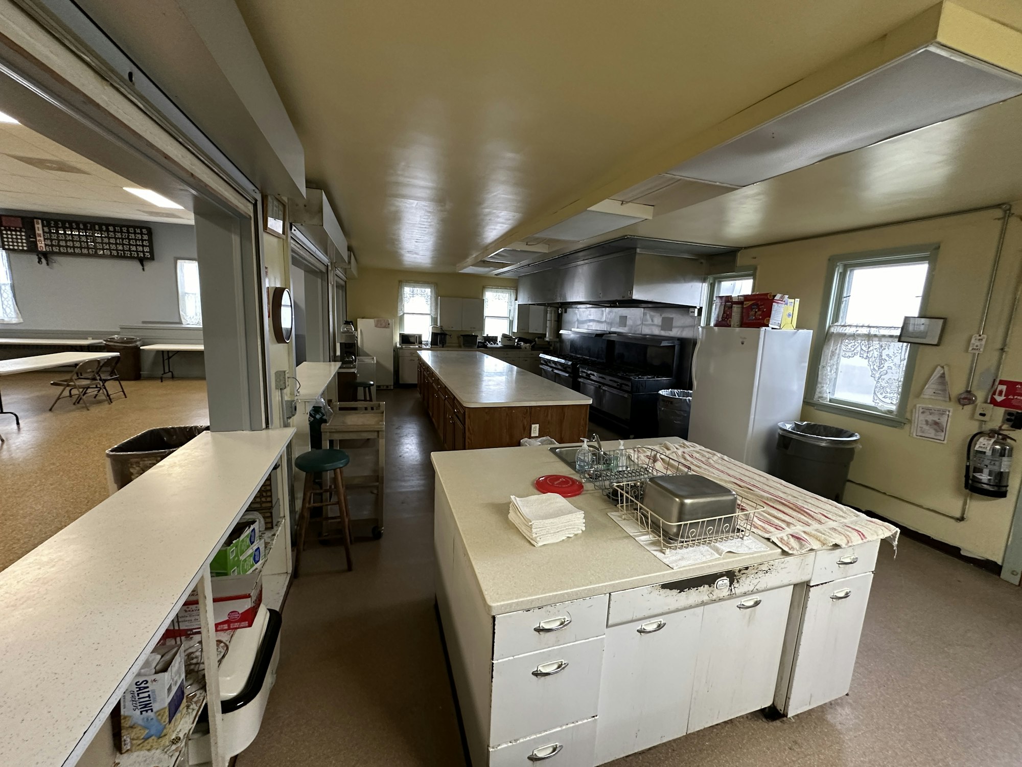 A spacious commercial kitchen with stainless steel appliances, counters, and a dining area with tables and chairs in the background.