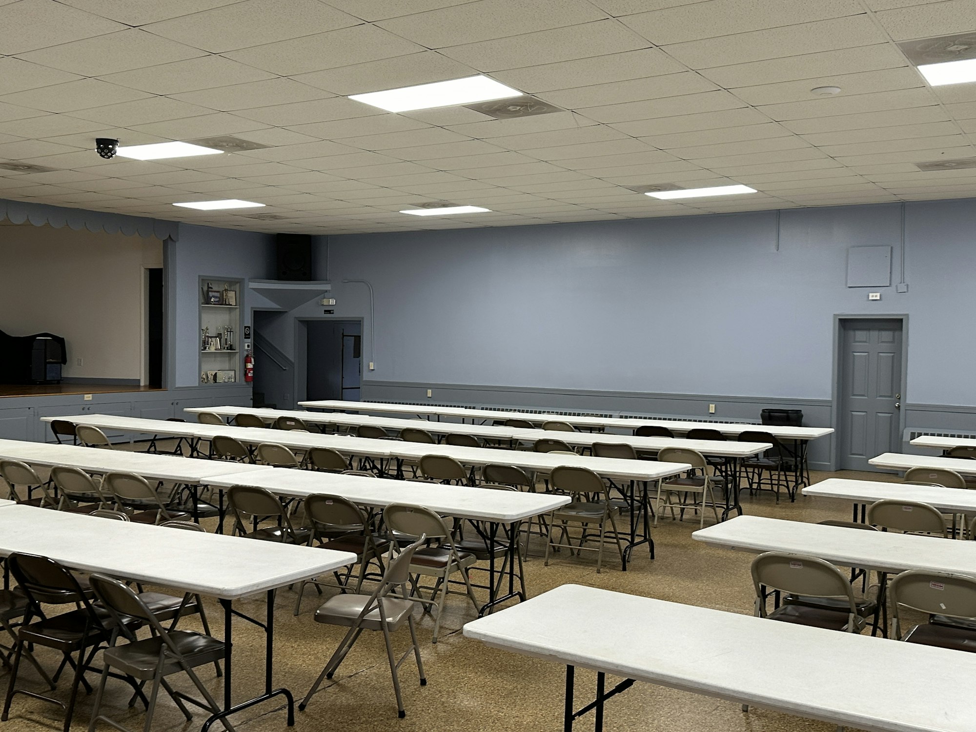 Empty meeting hall with rows of tables and folding chairs, a stage area, and a blue-gray color scheme.