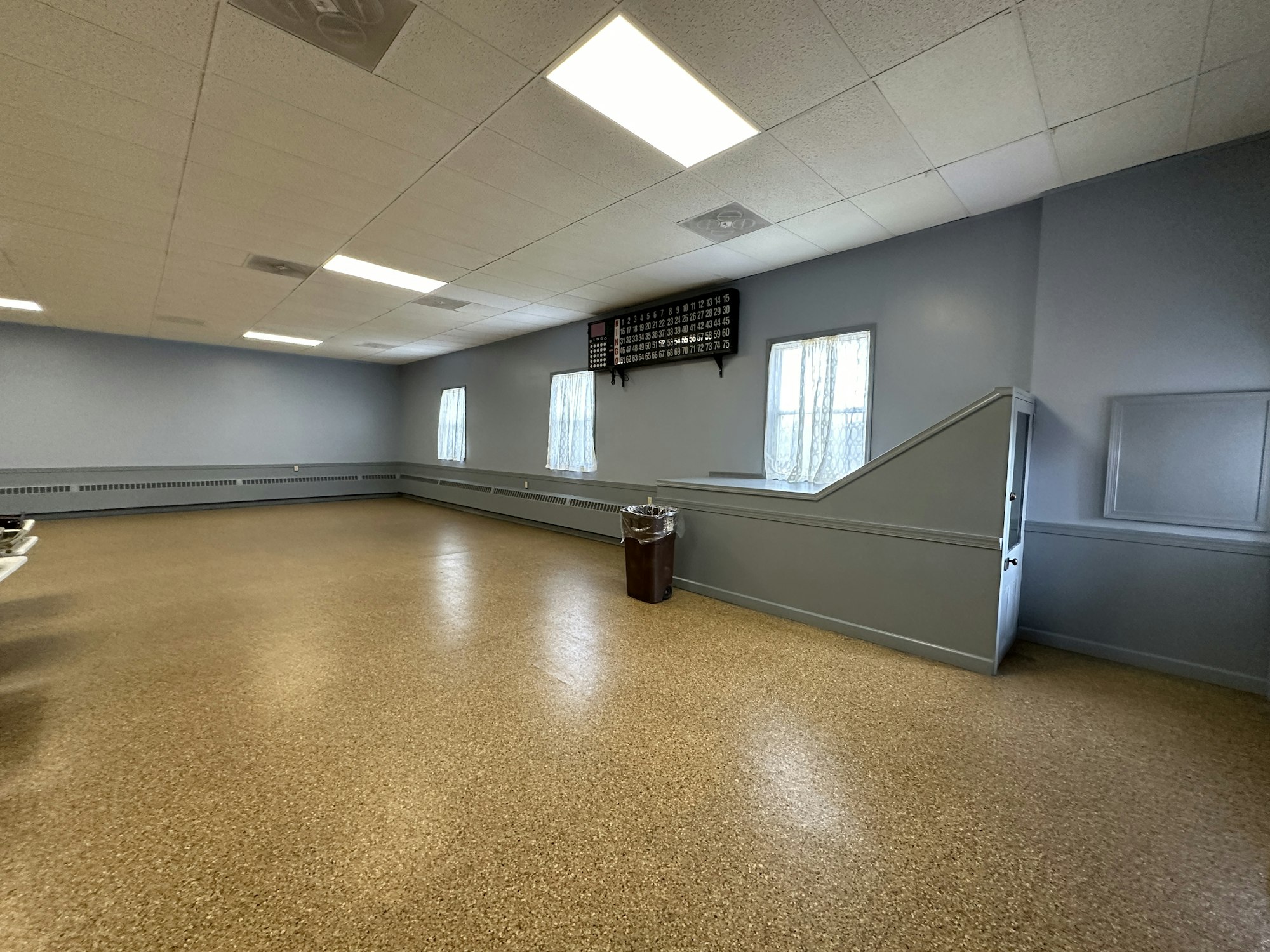 Empty room with cork floors, gray walls, fluorescent lights, a chair rail, windows with curtains, and a wall-mounted timetable/schedule.