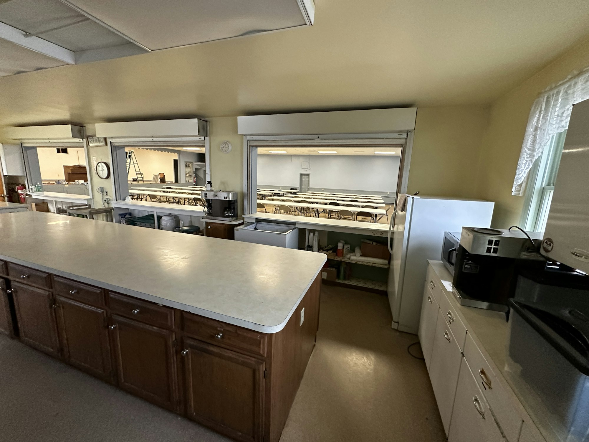 A kitchen with cabinets, appliances, and a view into a dining hall with tables and chairs.