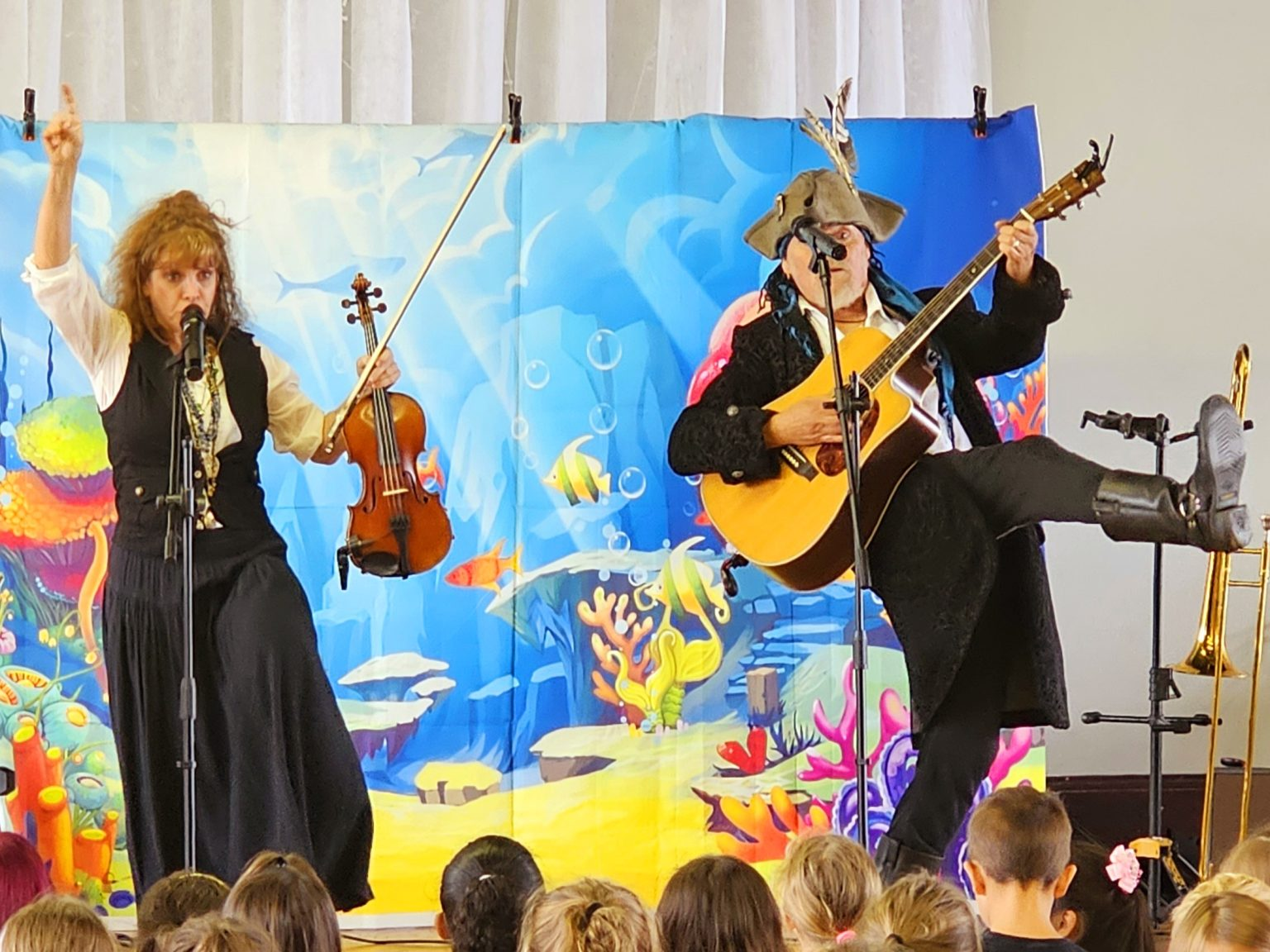 Two performers at a children's event, one with a violin and the other with a guitar, against an underwater-themed backdrop.