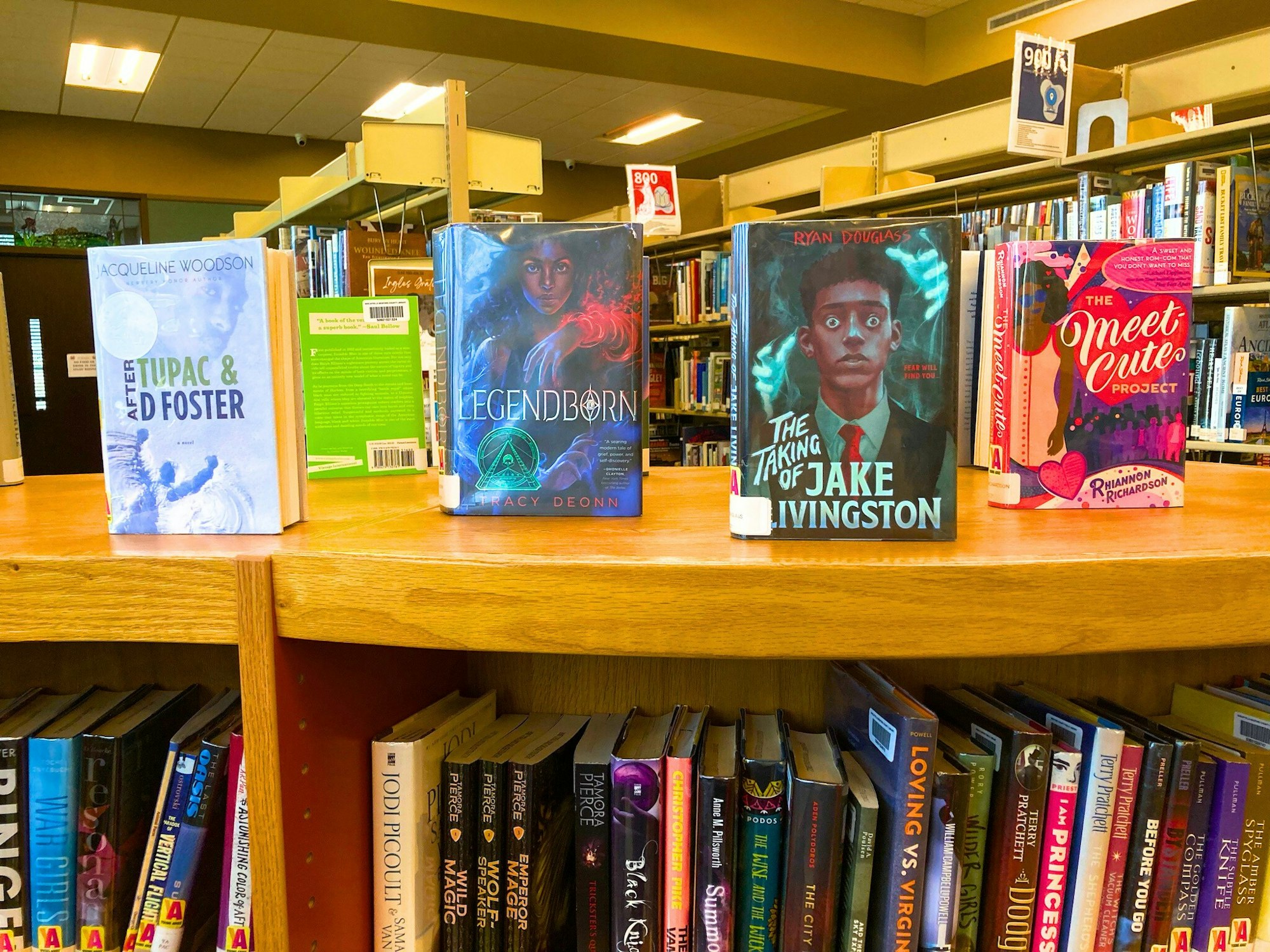 Books displayed on a library shelf: "After Tupac & D Foster," "Legendborn," "The Taking of Jake Livingston," "The Meet Cute Project."