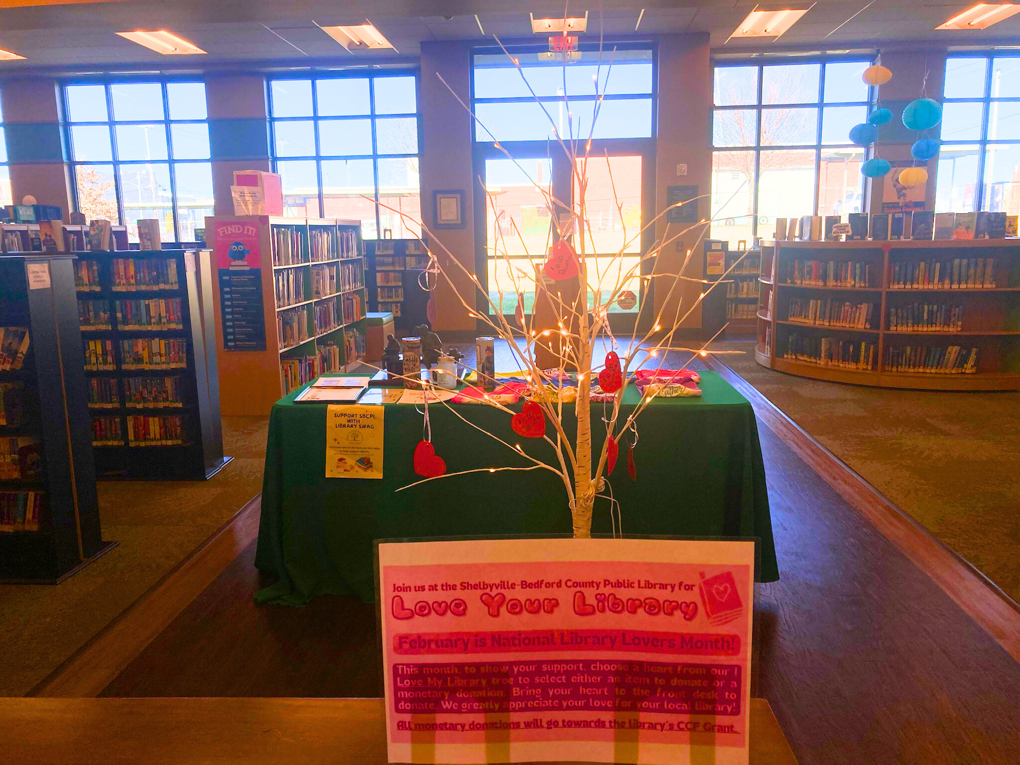 A library display for "Love Your Library" month with a decorated table, bookshelves, and signs promoting support for the library.