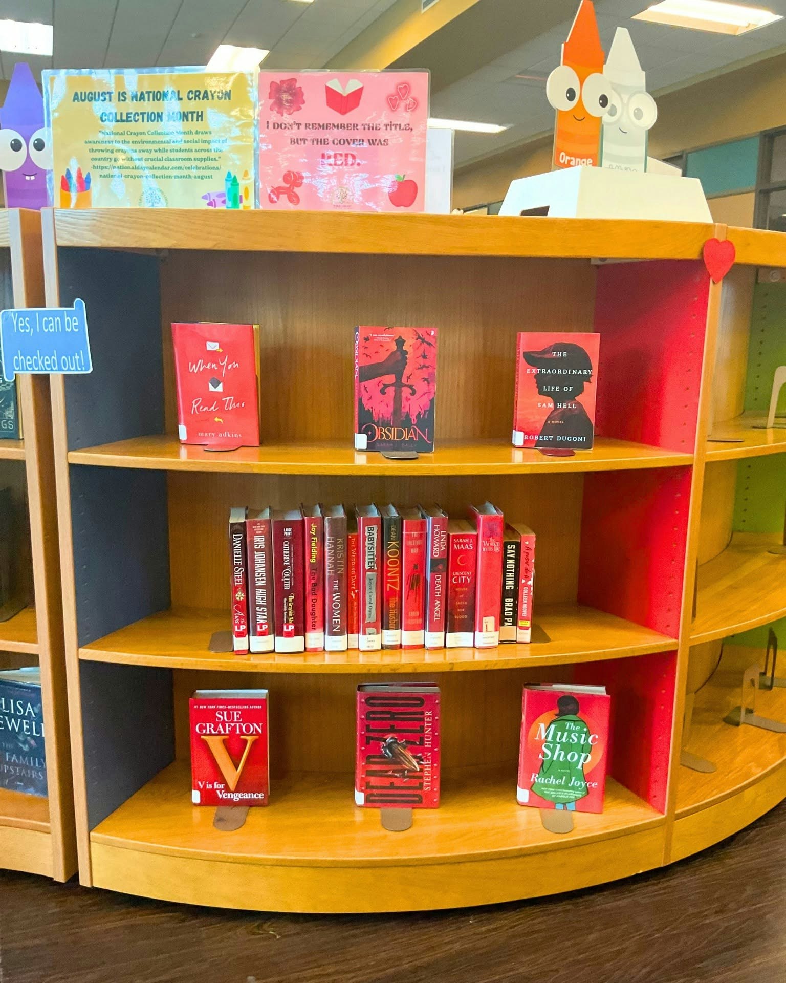 A library shelf featuring books with red covers, along with displays for National Crayon Collection Month and a playful sign.