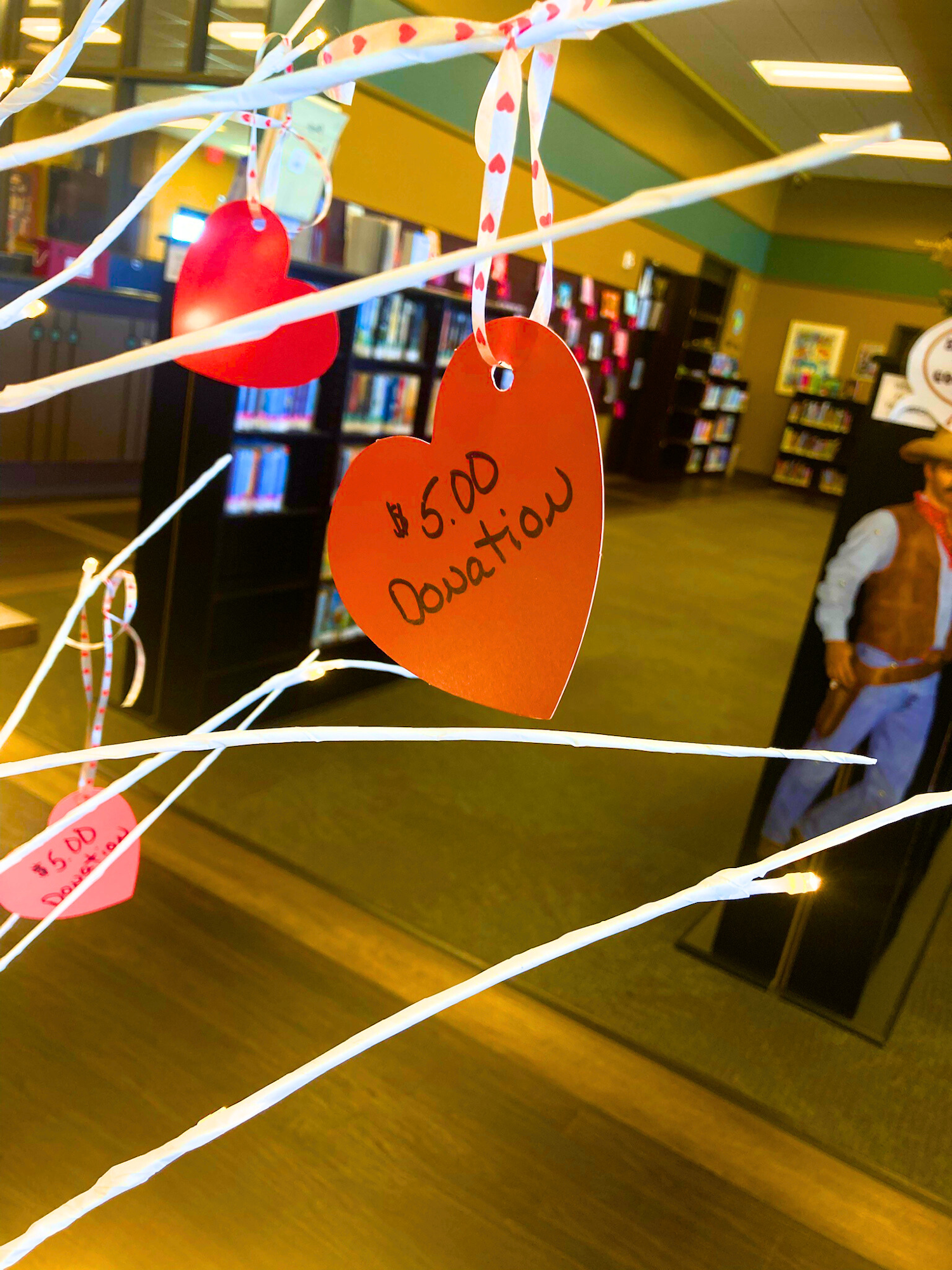 Paper hearts hang on a branch, each labeled "$5.00 Donation," in a library setting.