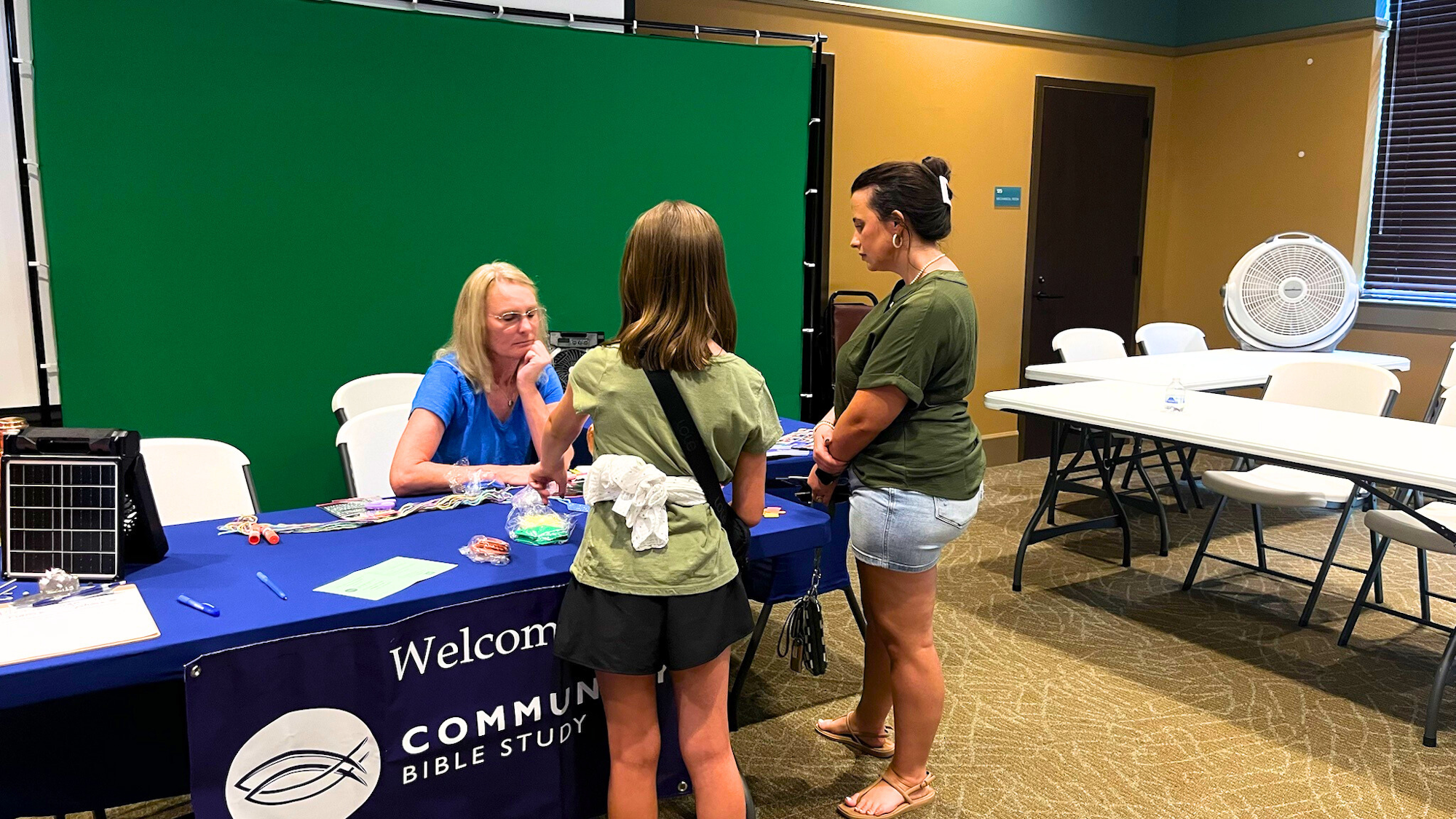 A woman and a child stand at a table talking to another woman. The table has a "Community Bible Study" banner.