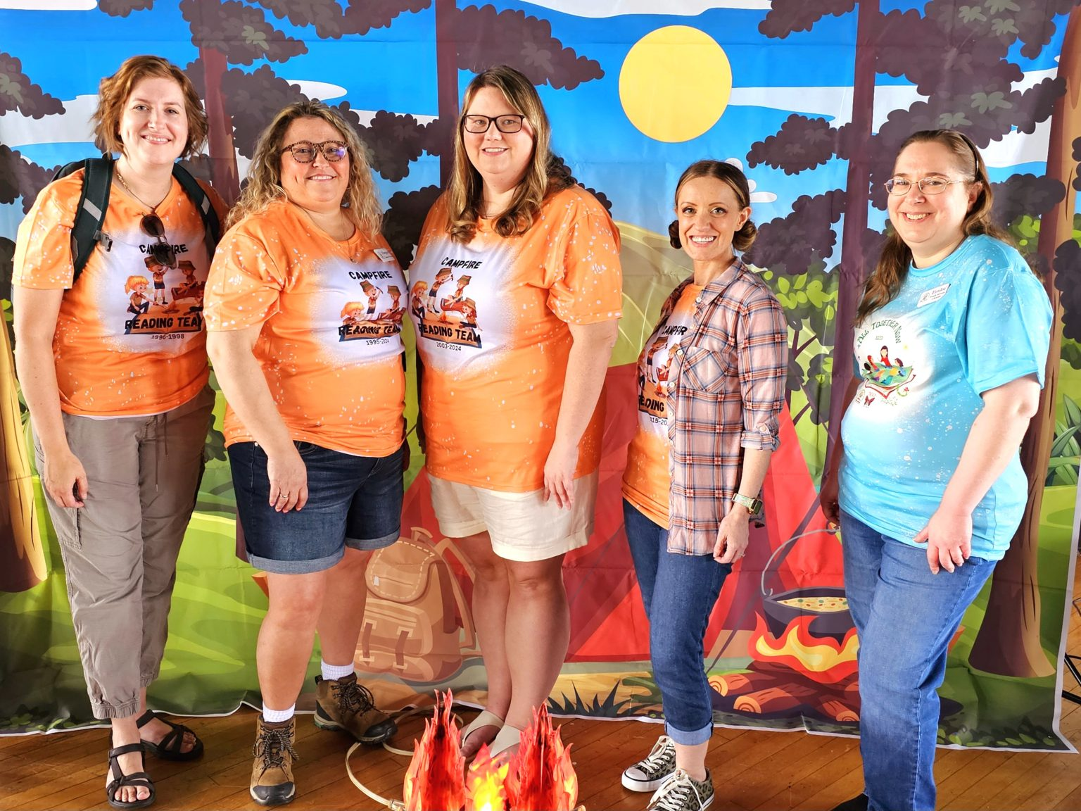 Five individuals with matching shirts posing in front of a campfire-themed backdrop.
