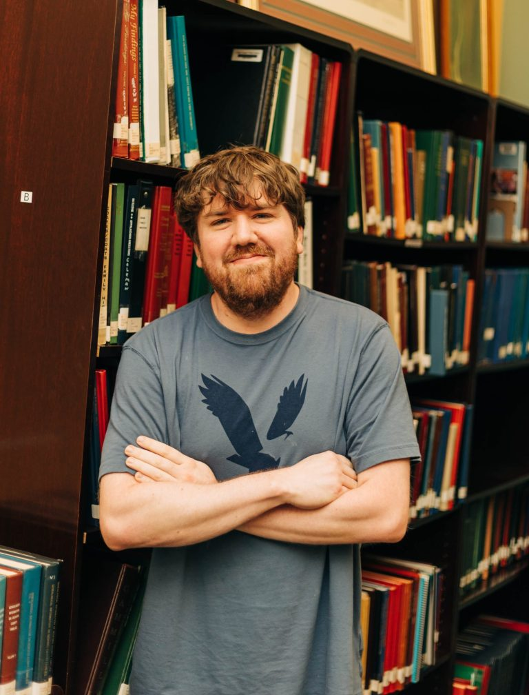 A man with arms crossed standing in front of a bookshelf.
