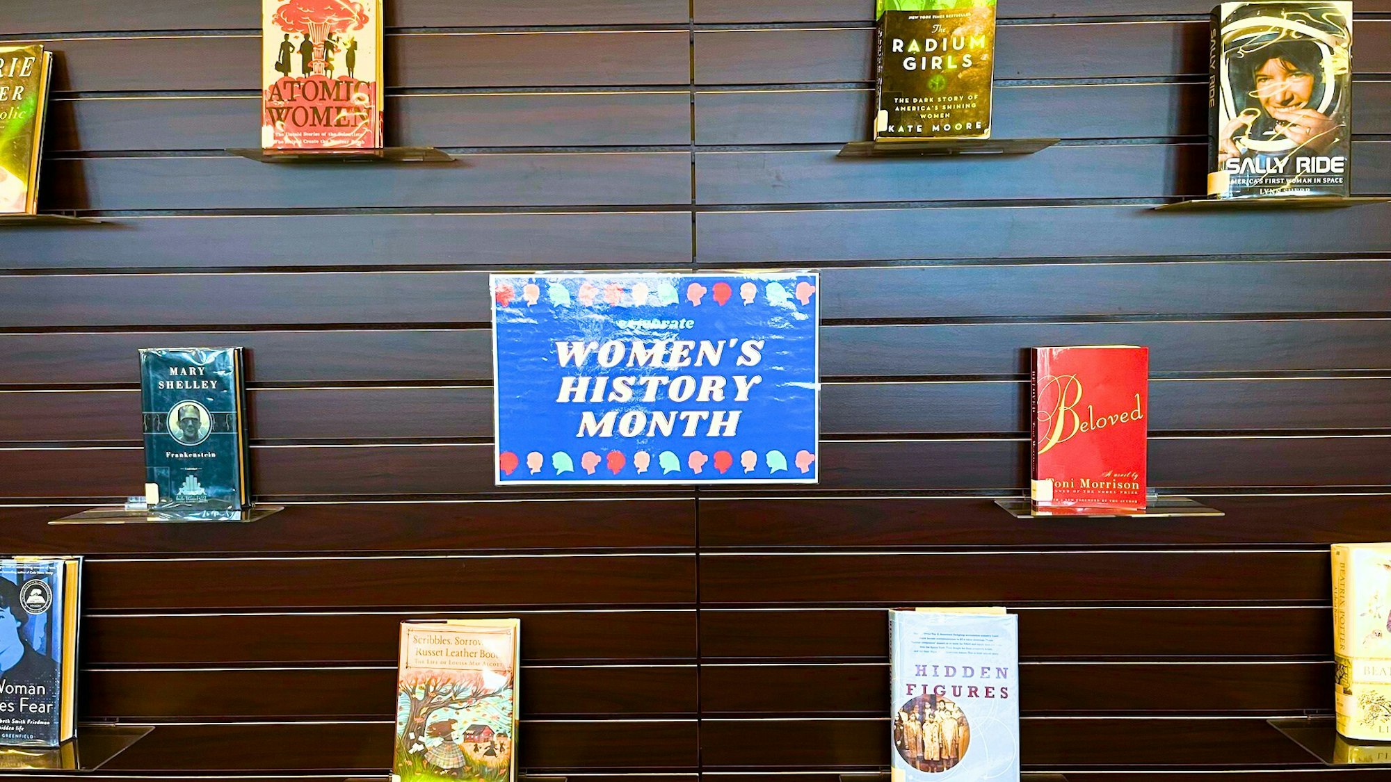 Books displayed on a wall with a "Women's History Month" sign in the center.