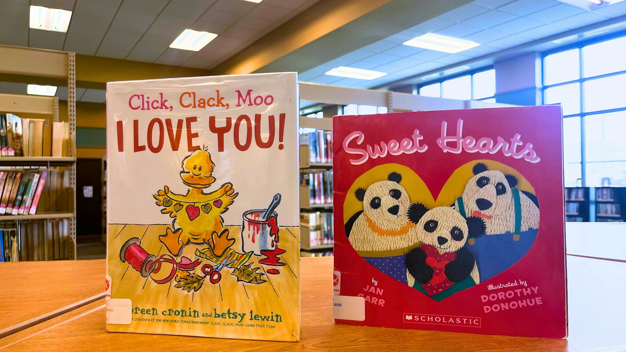 Two children's books on a library table: "Click, Clack, Moo I Love You!" and "Sweet Hearts" featuring panda illustrations.