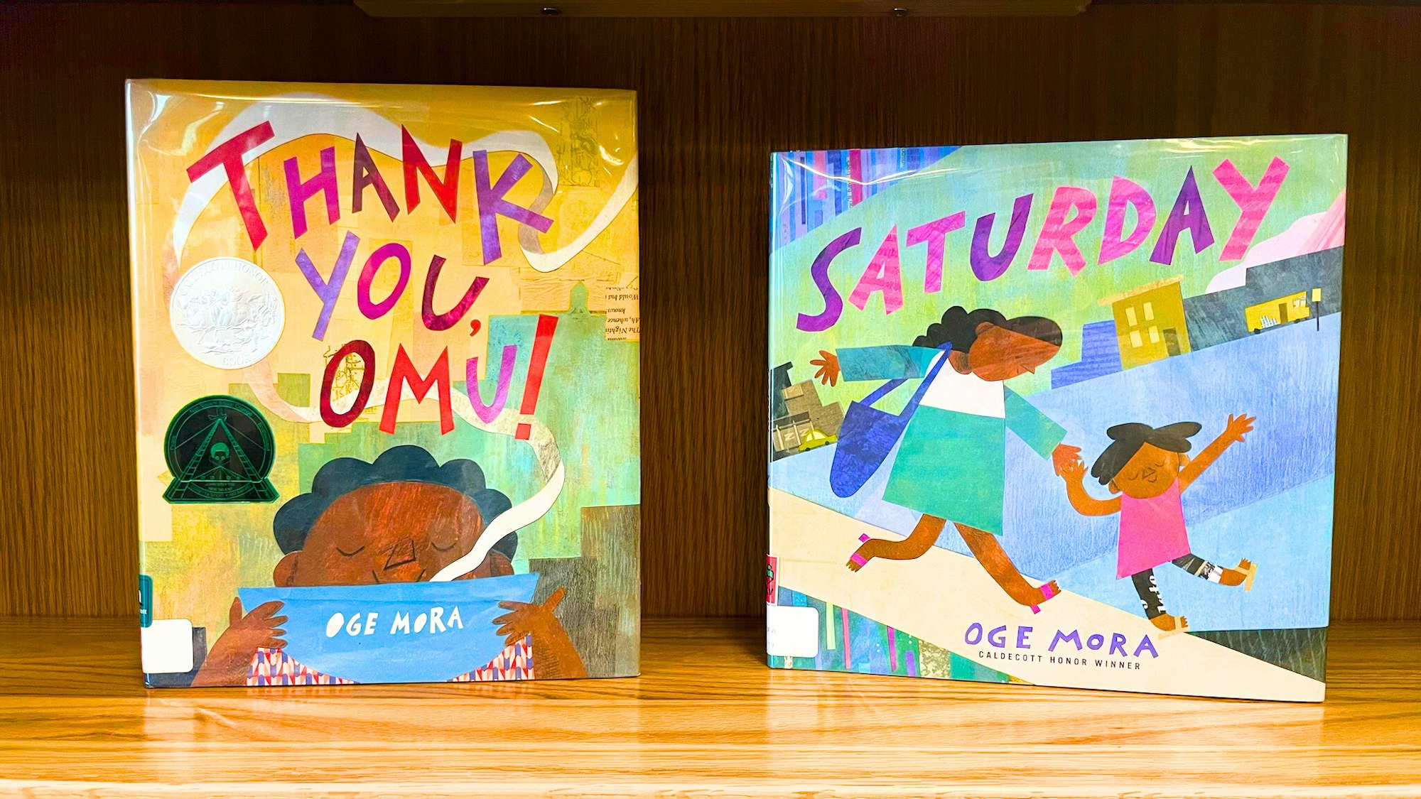 Two children's books by Oge Mora, titled "Thank You, Omu!" and "Saturday," displayed on a wooden shelf.