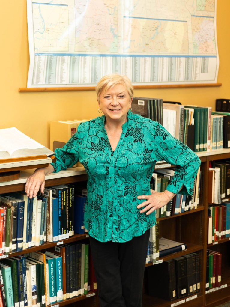 A smiling woman standing in a library with books and a map in the background.