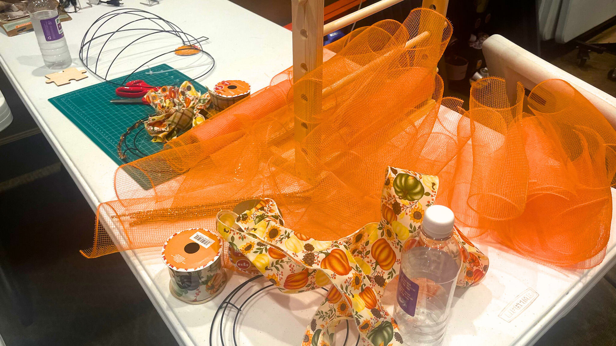 Crafting table with orange mesh, floral ribbons, wire frames, scissors, cutting mat, and a water bottle.