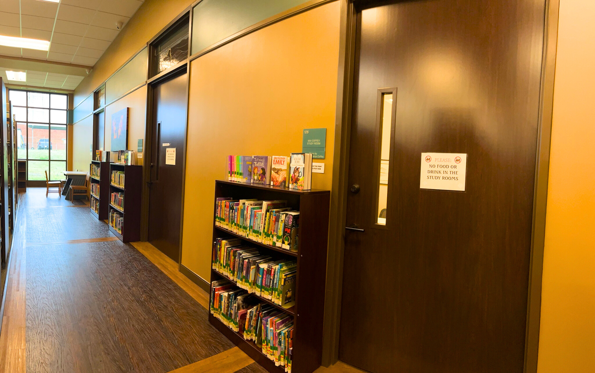 Library hallway with wooden shelves of books, a door labeled "Study Room," and a sign prohibiting food and drink.