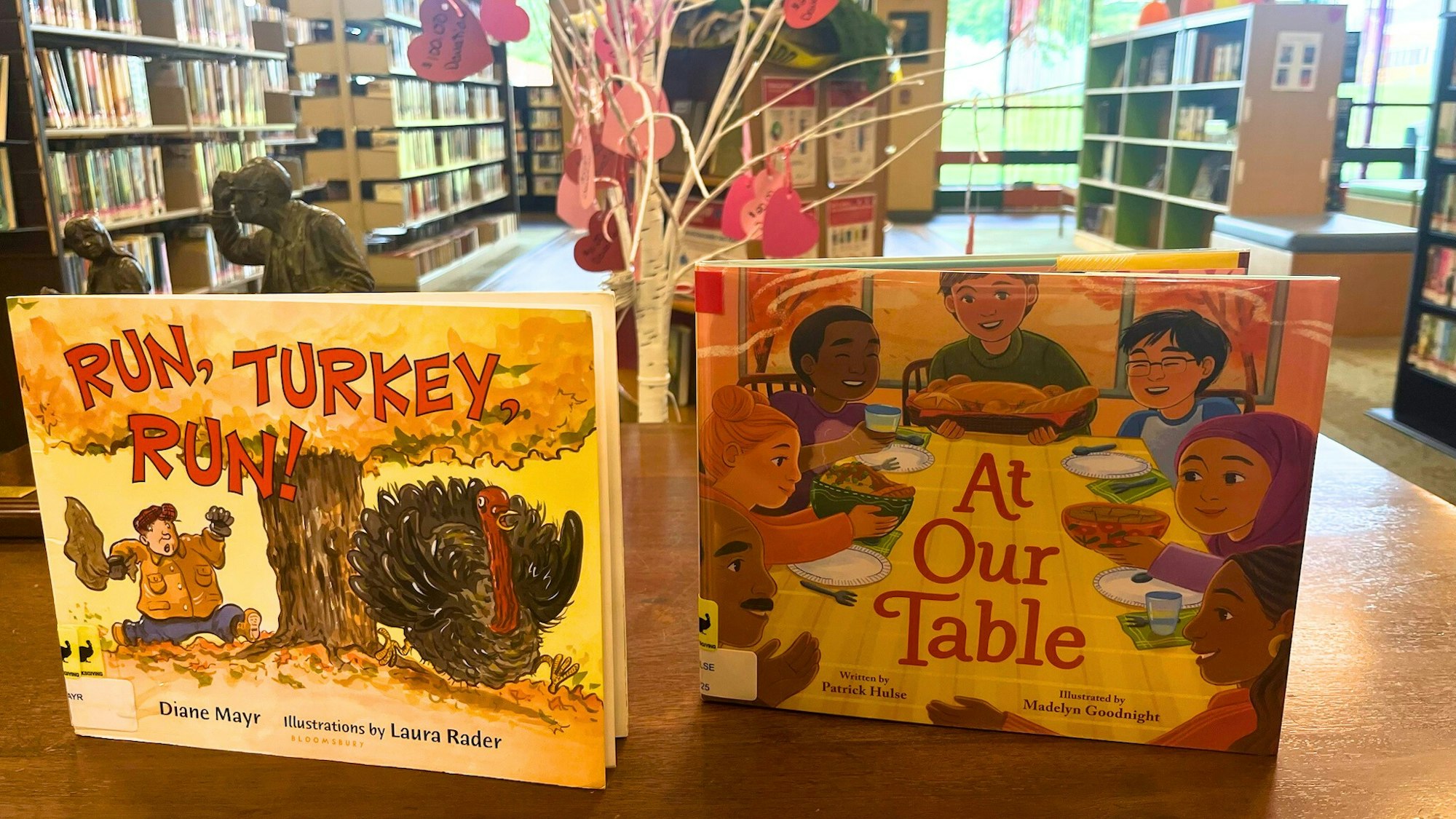 A library scene featuring two children's books: "Run, Turkey, Run!" and "At Our Table," illustrating themes of Thanksgiving and community.