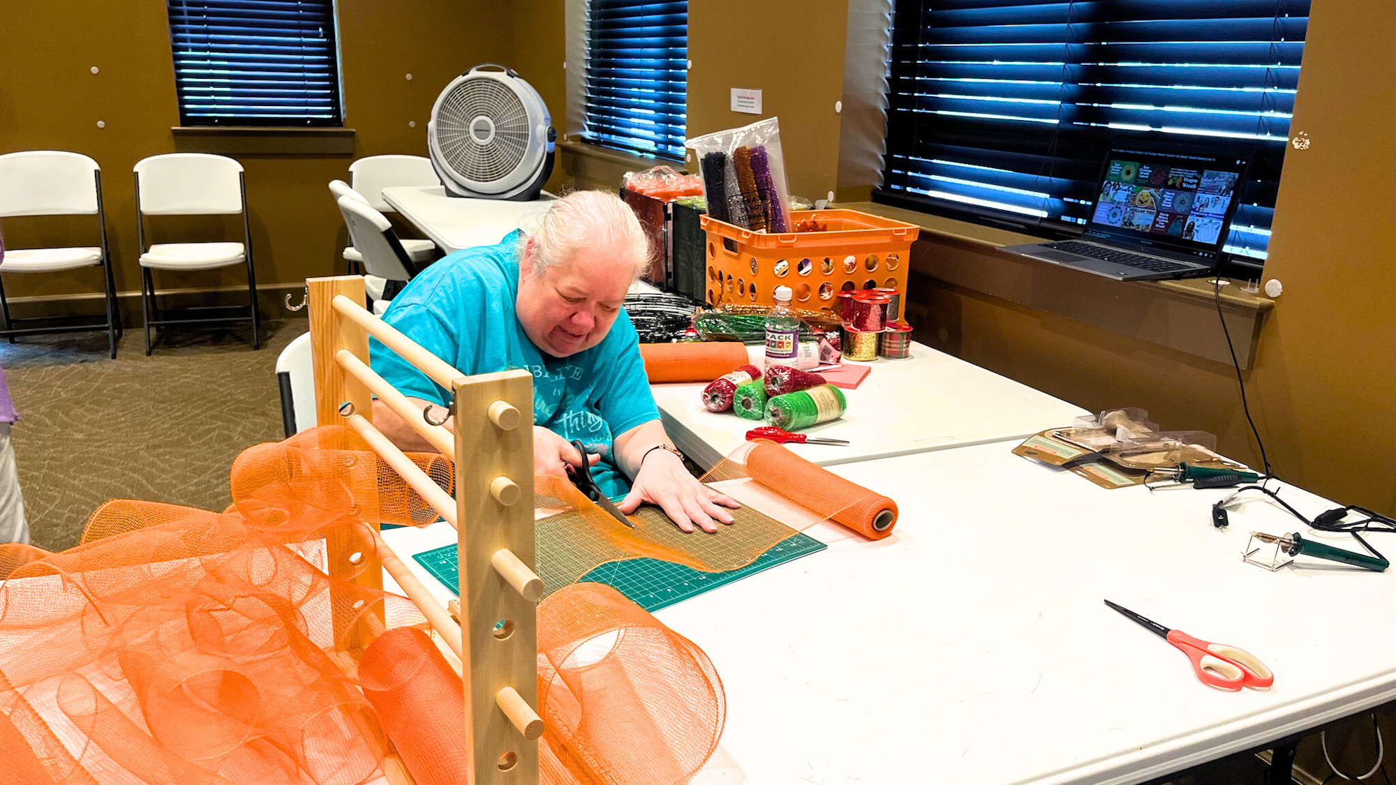 A person cuts orange mesh on a table with craft supplies and tools; a laptop and fan are in the background.