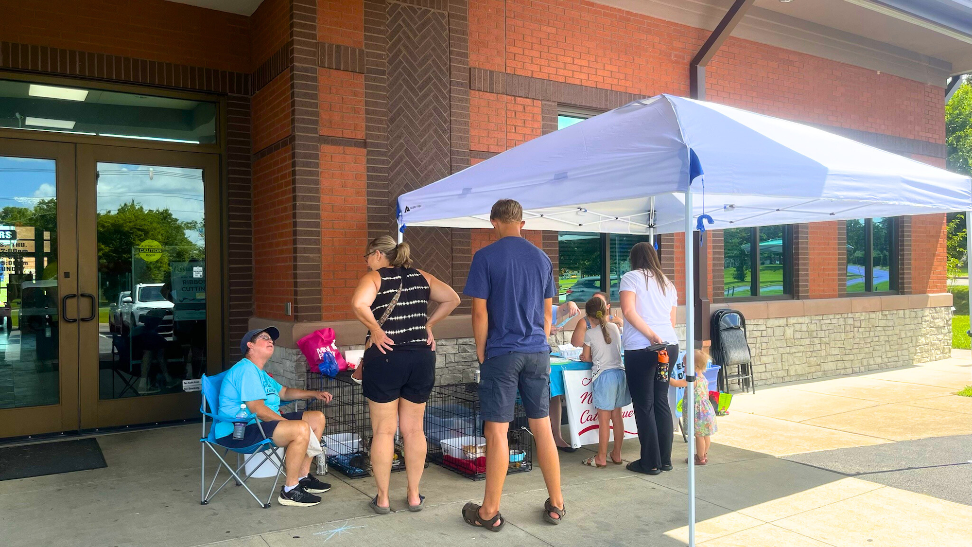 A group of people gather under a tent outside a building, likely for an event or sale, with pets in cages nearby.