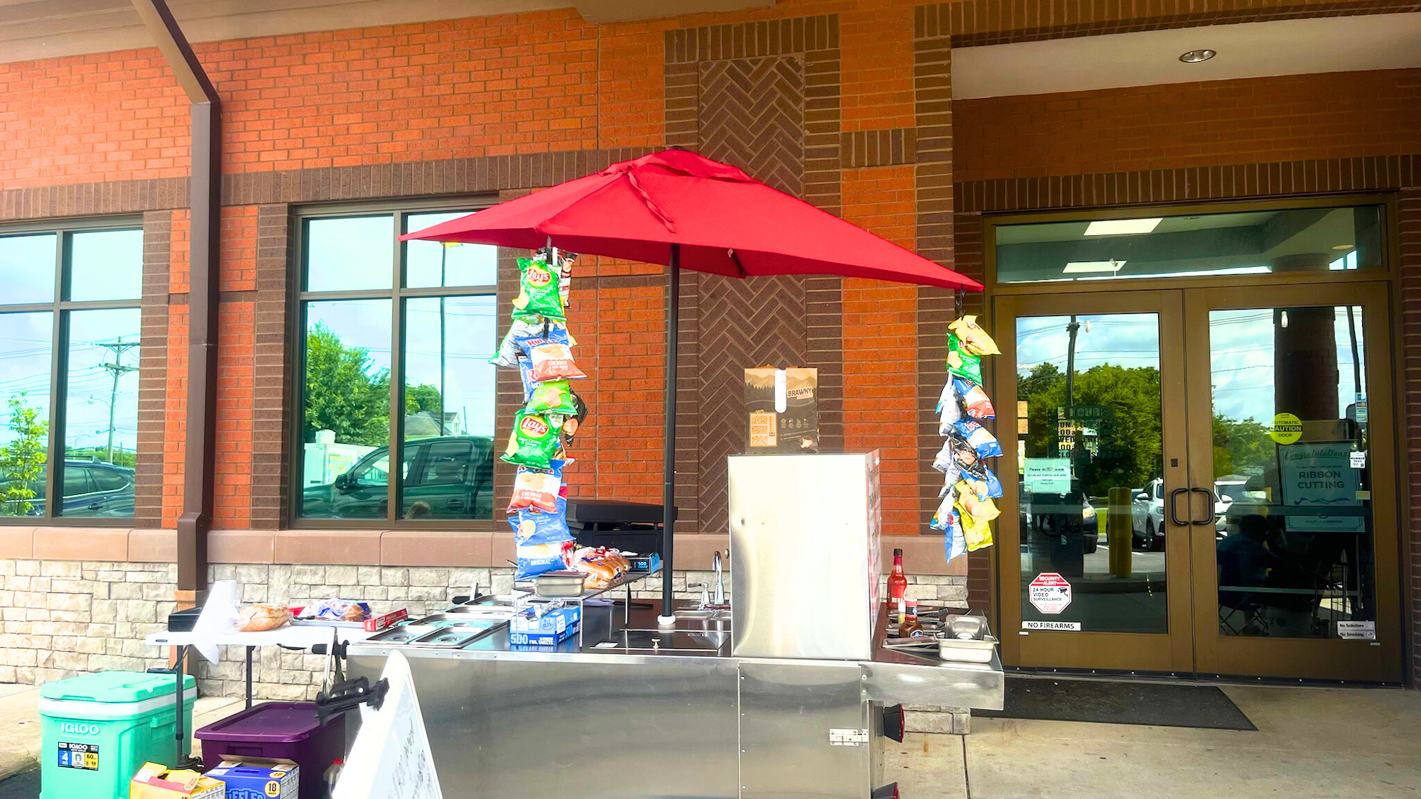 Snack stand with chips and a red umbrella outside a building entrance.
