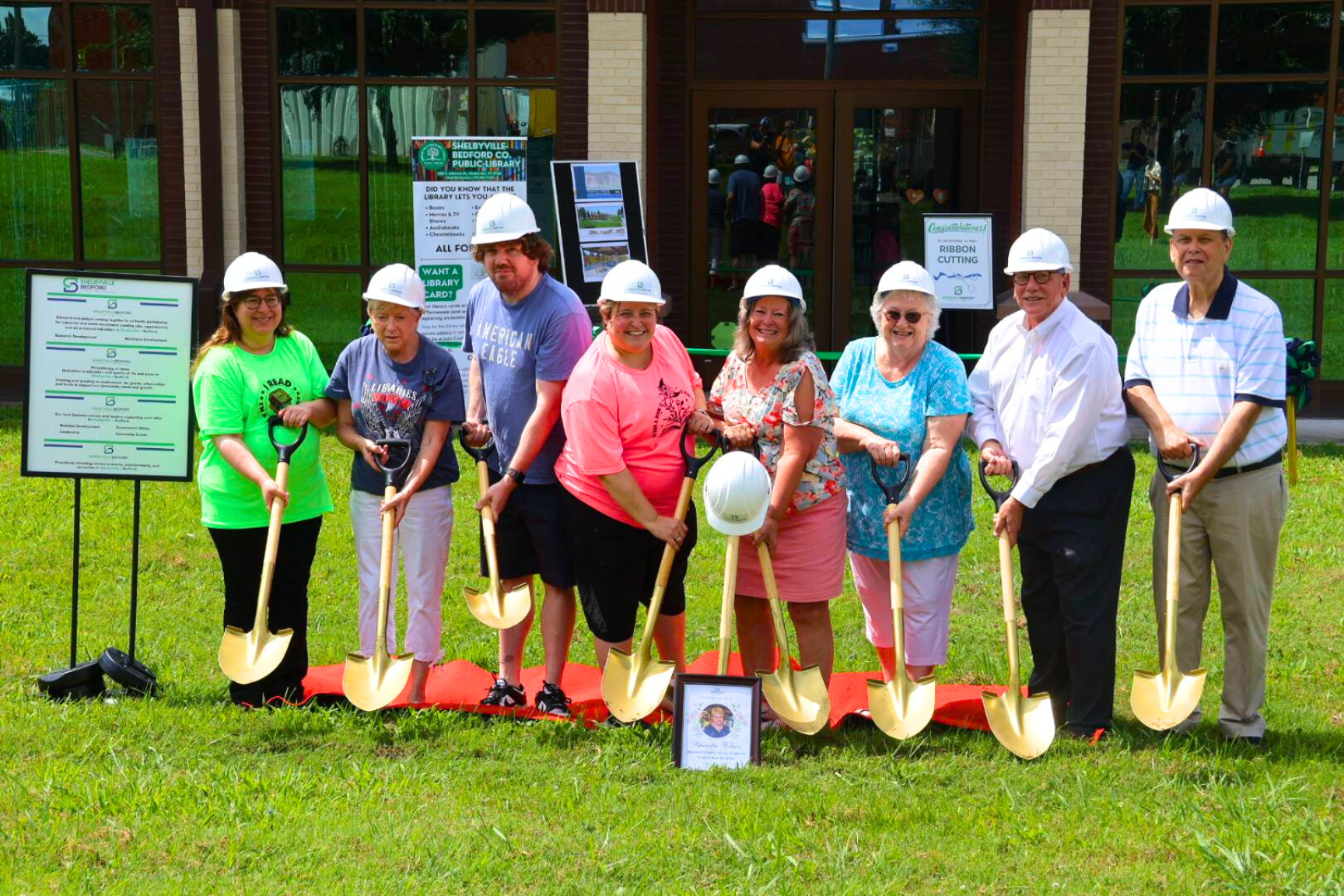A group of people in hard hats hold shovels, posing for a groundbreaking ceremony outside a building.