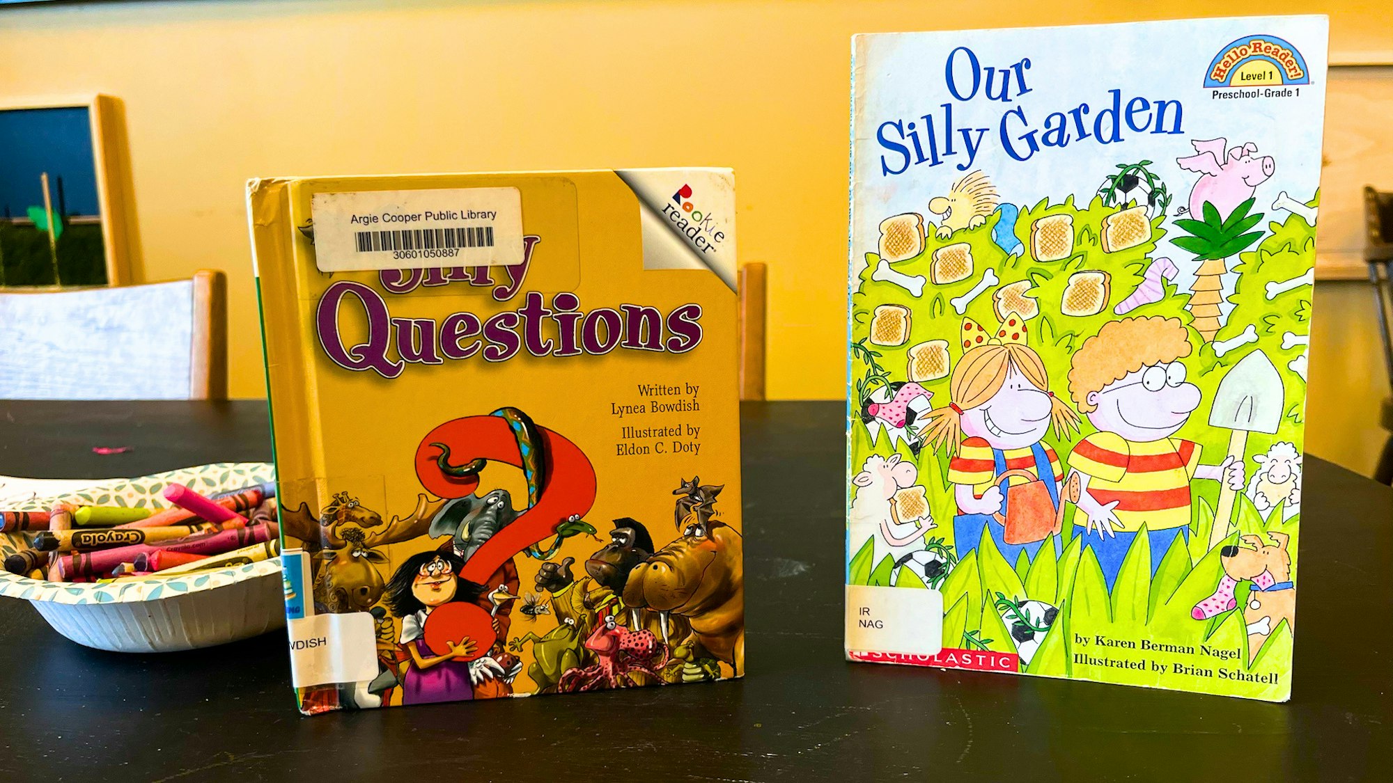 Two children’s books: "Silly Questions" and "Our Silly Garden," alongside a bowl of crayons on a table.
