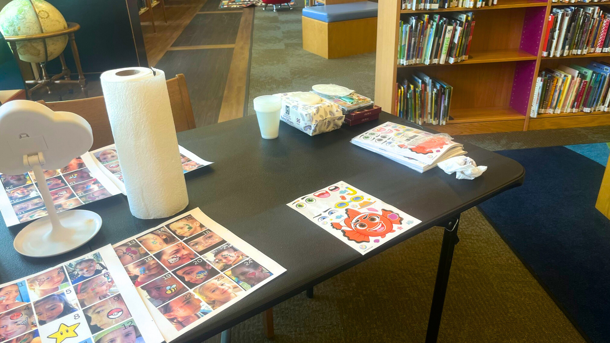 A table with face painting options, paper towels, tissue box, plastic cups, and library shelves in the background.