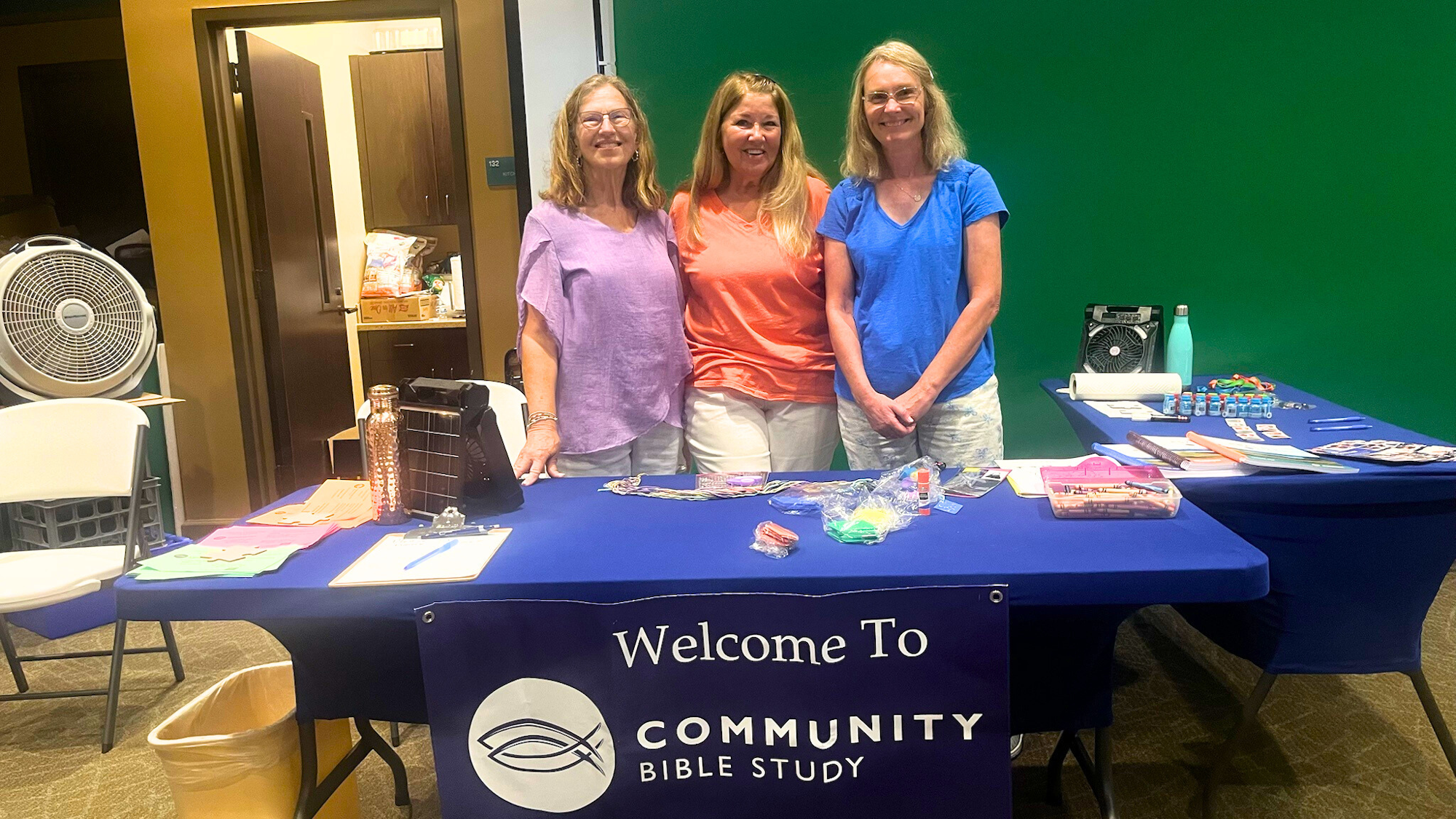 Three people at a booth with a "Welcome to Community Bible Study" sign, pamphlets, and supplies on the table.