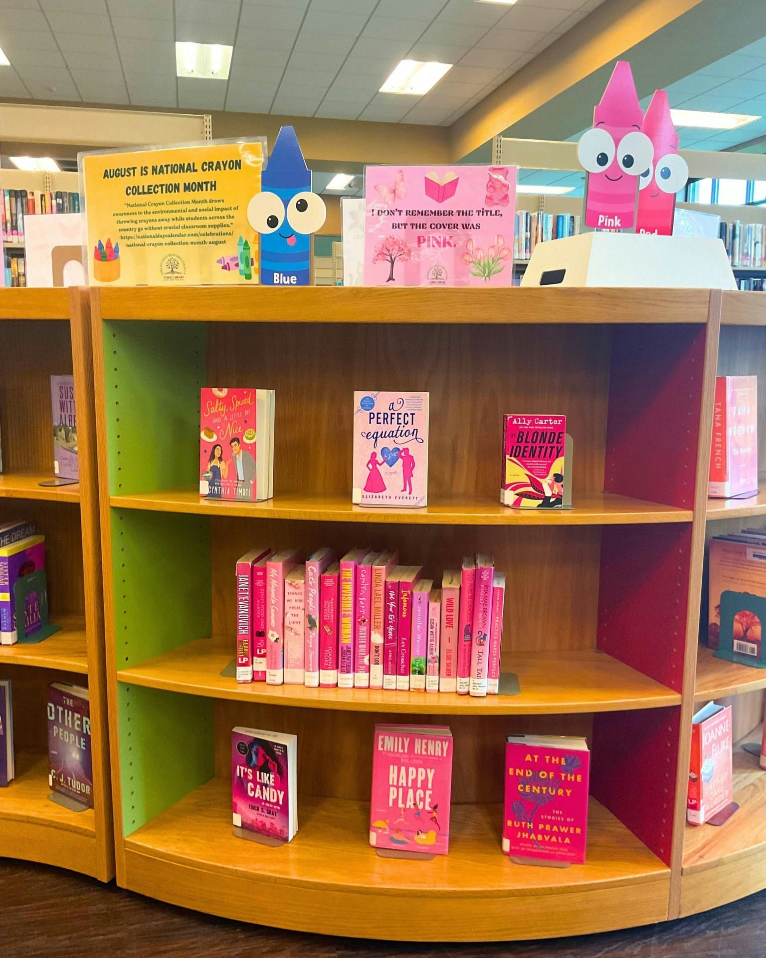 A library shelf displays pink-themed books for National Crayon Collection Month, featuring colorful crayon decorations and signage.