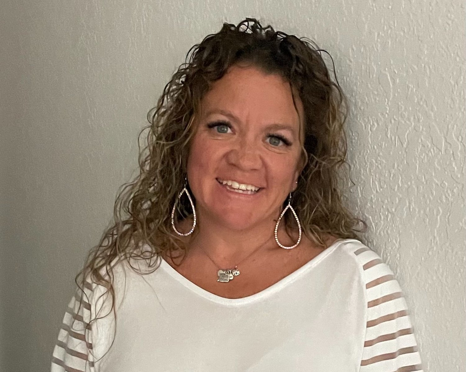 A smiling woman with curly hair, wearing a white top and large earrings, standing against a textured wall.
