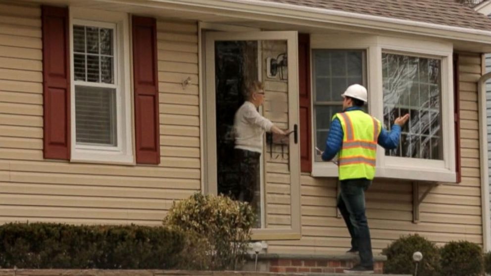 A person in a high-visibility vest and helmet speaks to another at their front door.