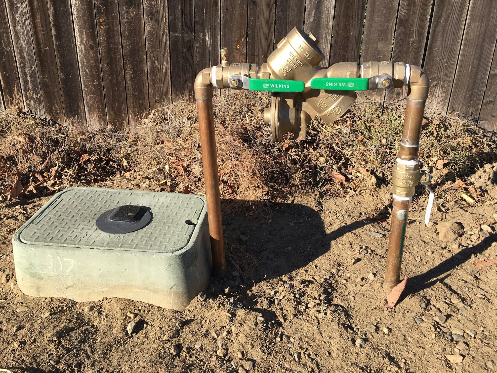 The image shows a water meter setup with pipes, a shut-off valve, and a protective green cover in a dirt area by a wooden fence.
