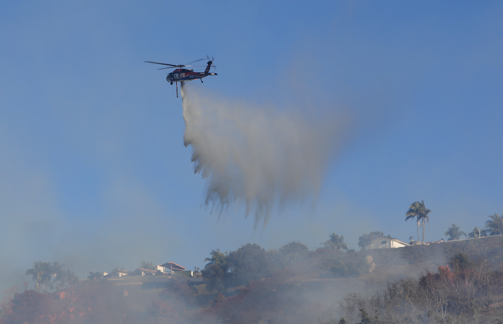 Un helicóptero lanza agua sobre una humeante ladera con casas en el área de servicio de Rainbow.