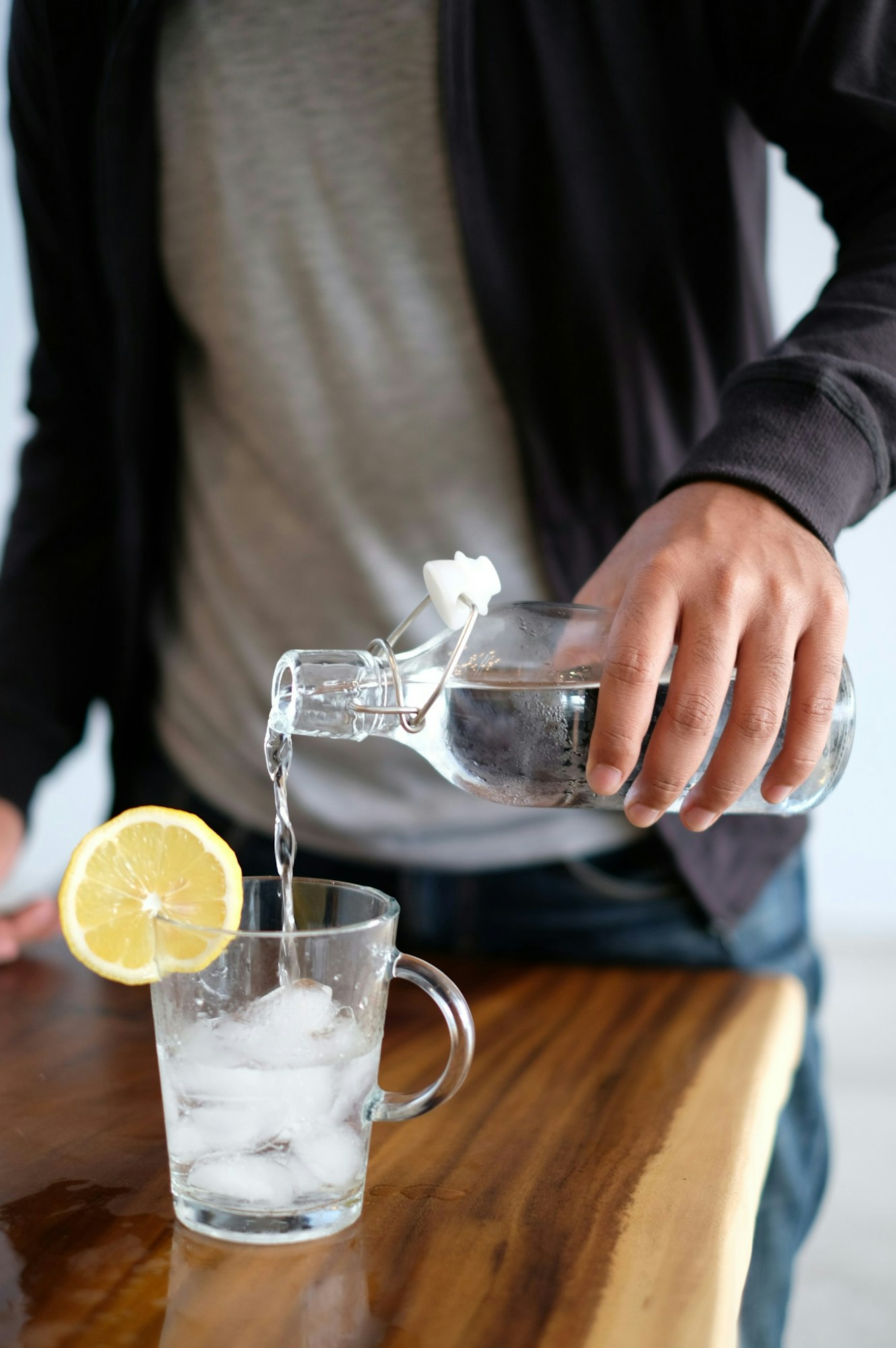 A person pouring water into a glass with ice cubes, lemon slice on the counter.