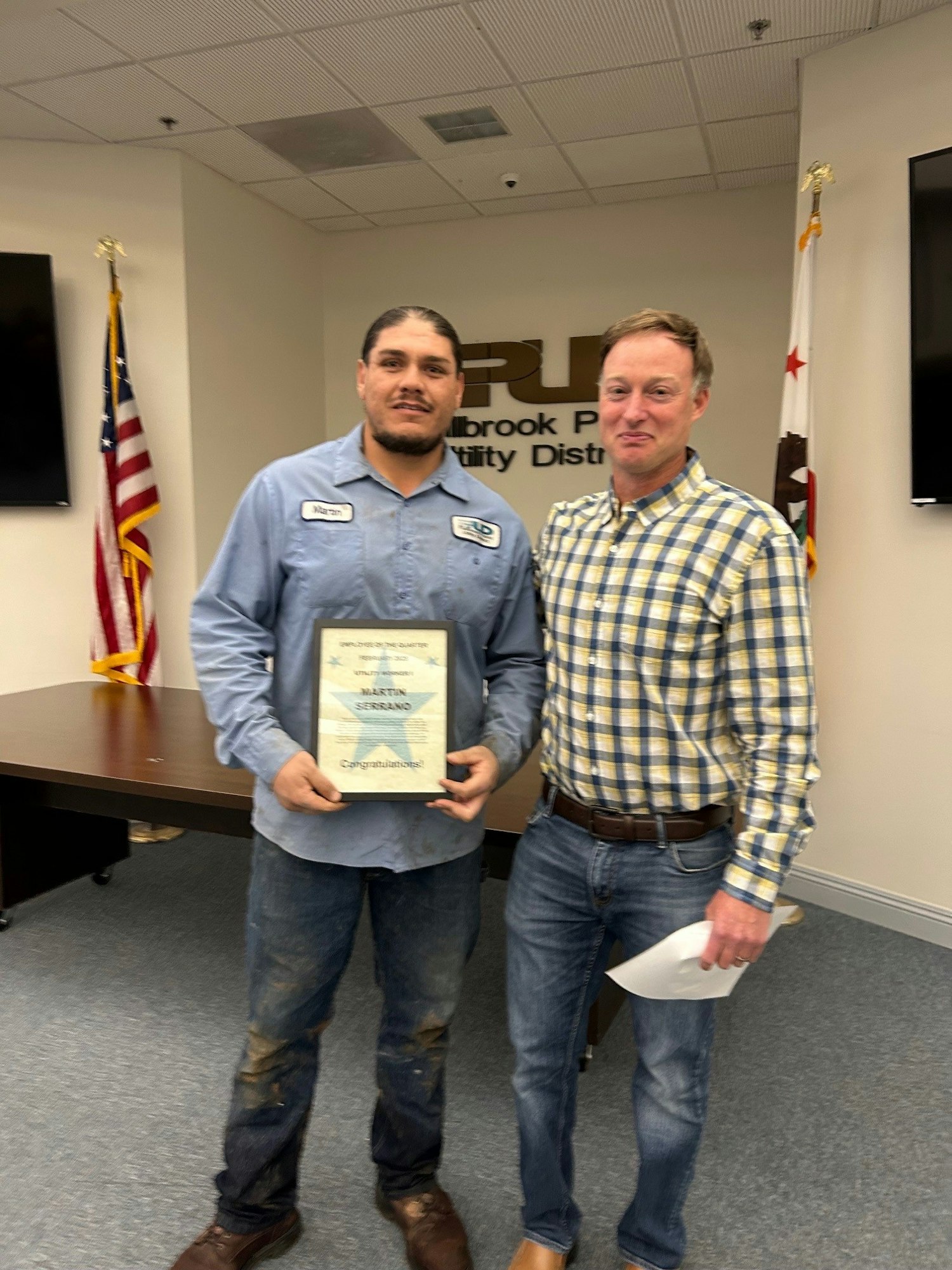 Two men pose for a photo in an office setting, one holding an employee recognition certificate, with flags in the background.