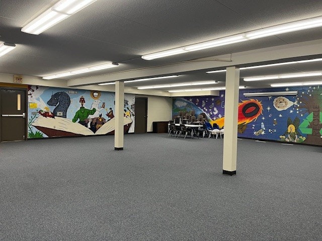 The image shows a spacious room with a carpeted floor, featuring colorful murals on the walls and a few chairs stacked.
