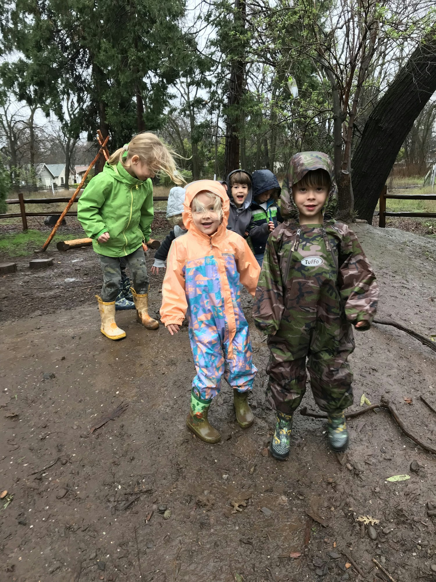 Kids in raincoats and boots standing on a muddy path in a wooded area.