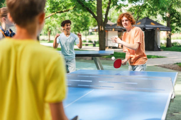 A group of kids playing table tennis outdoors, enjoying a lively game in a green park setting.