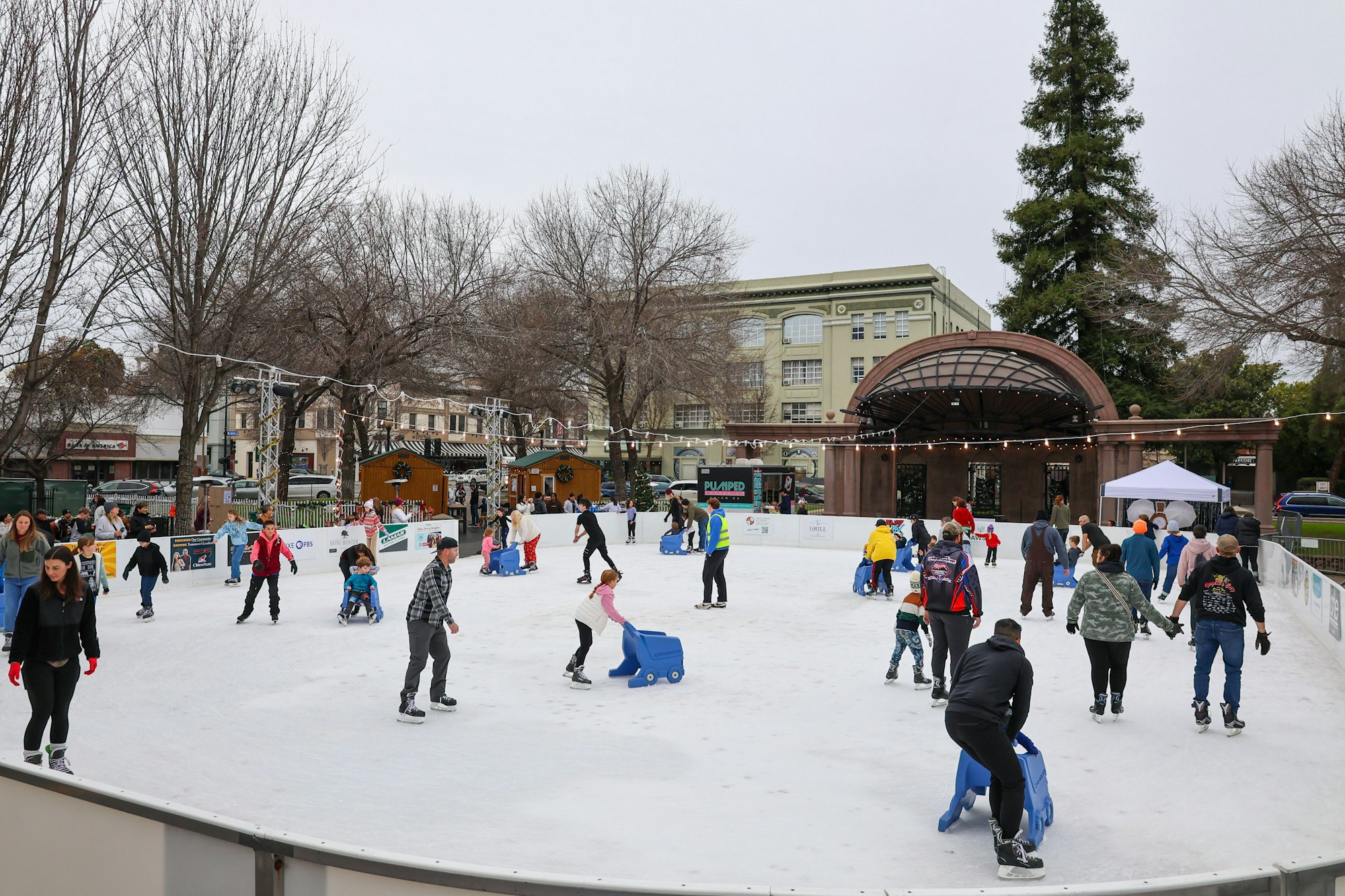 People ice skating in an outdoor rink, some with blue skate aids, surrounded by buildings and trees.